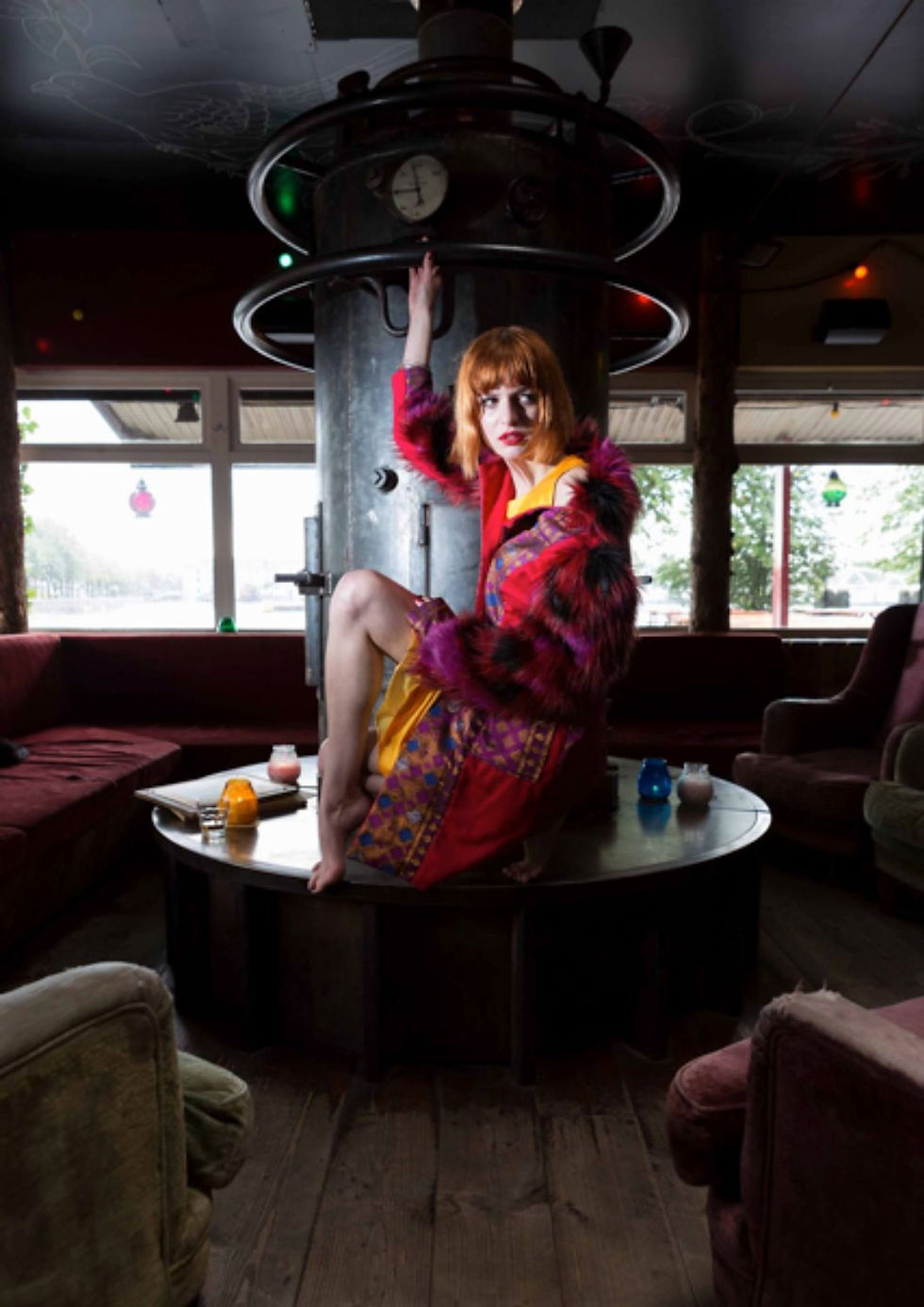 A person in a colorful outfit poses on a round table in a dim room with plush seating. They're surrounded by candles and glassware.