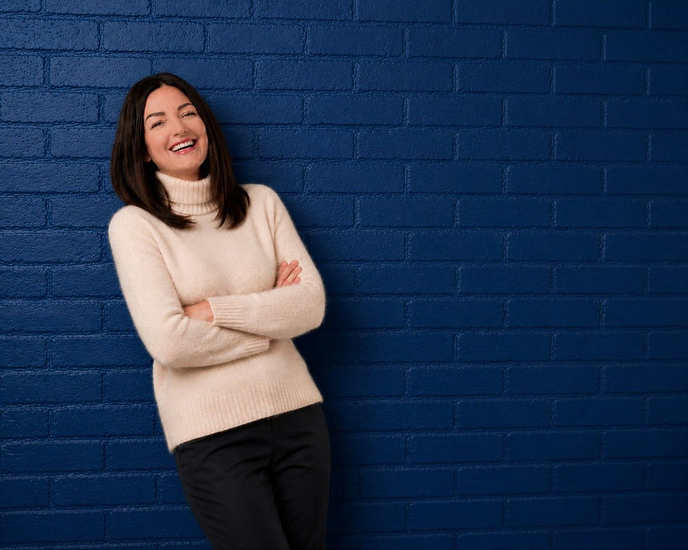Woman in a cream sweater leans on a blue brick wall, arms crossed and smiling warmly. She exudes a relaxed, cheerful mood.