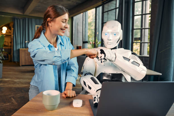 Woman in a blue shirt fist-bumps a robot in a cozy room with large windows. A mug and laptop are on the table, creating a friendly mood.