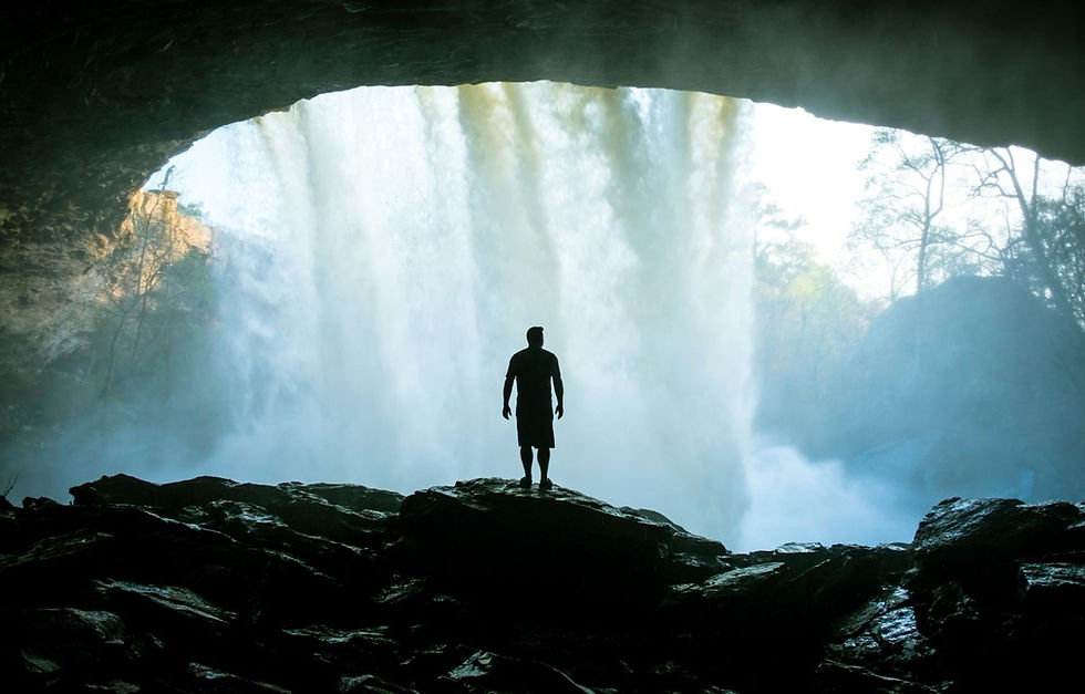 Silhouette of a person standing on rocks in a cave, overlooking a waterfall with misty blue and green hues in the background.