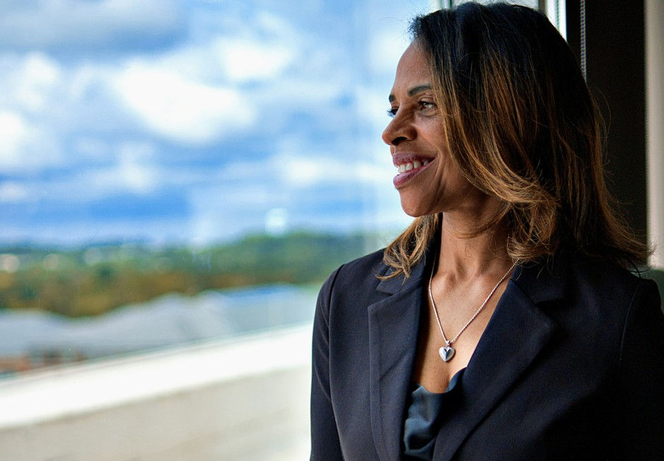 Smiling woman in black blazer and heart necklace looks out a window at a cloudy sky and distant landscape.