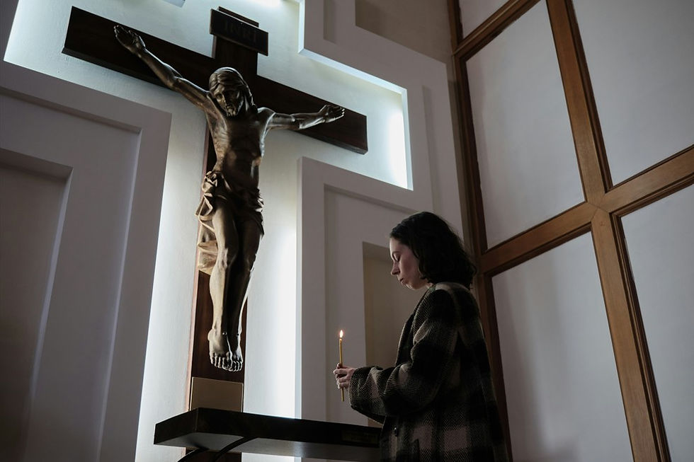 Woman holds a lit candle before a large crucifix in a quiet chapel with white walls and wooden panels.