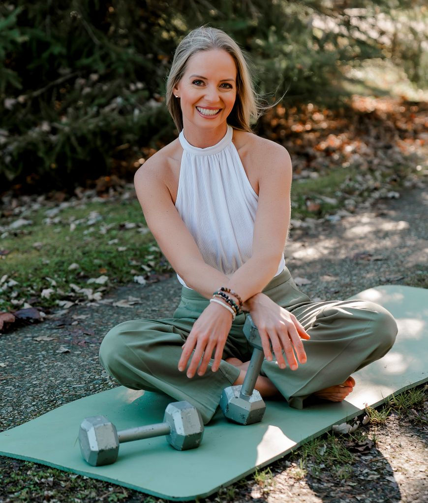 Smiling woman sits cross-legged on a yoga mat outdoors, hands on dumbbells. She's wearing a white top and green pants, surrounded by greenery.