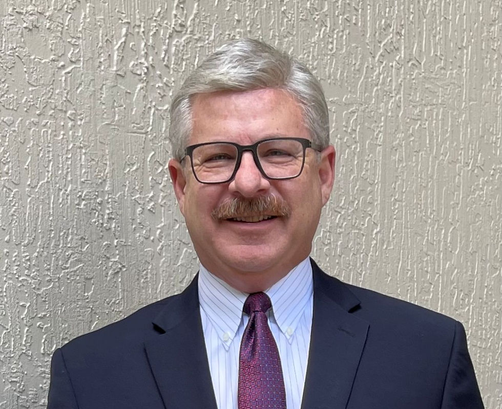 Smiling man with glasses in a suit and tie stands against a textured beige wall, conveying a professional and friendly demeanor.