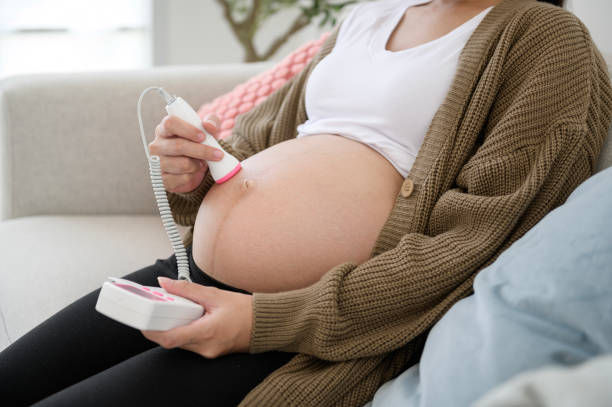 Pregnant woman in brown cardigan using a pink fetal Doppler on her belly, sitting on a beige sofa. Cozy home setting.