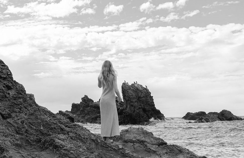 a woman standing on rocky terrain near the ocean, gazing at the distant rocks and waves.