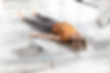 Woman in orange shirt and gray leggings lies on yoga mat, eyes closed, beside an open laptop on a wooden floor, creating a calm mood.