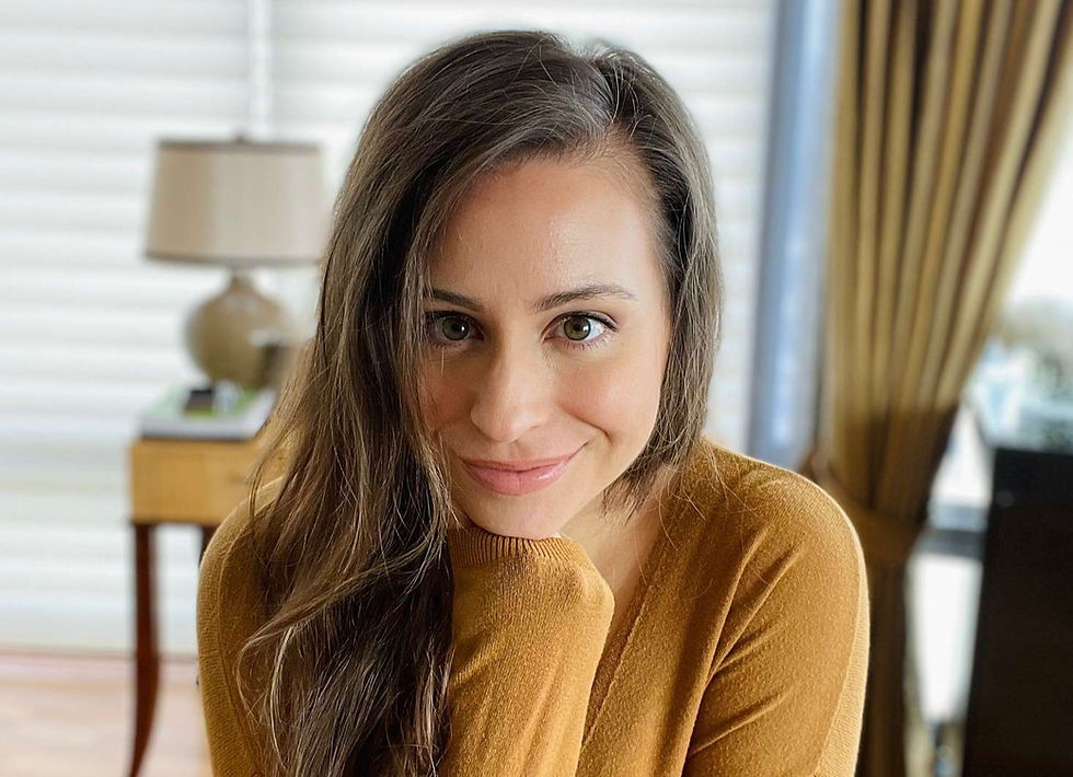 Christina Giordano with long hair in mustard sweater, leaning on hand. Room with soft lighting, lamp, and curtains in the blurred background.