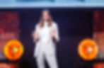 A photo of Kerdu in a light suit stands on stage, smiling and gesturing with both hands raised, framed by two bright orange stage lights.