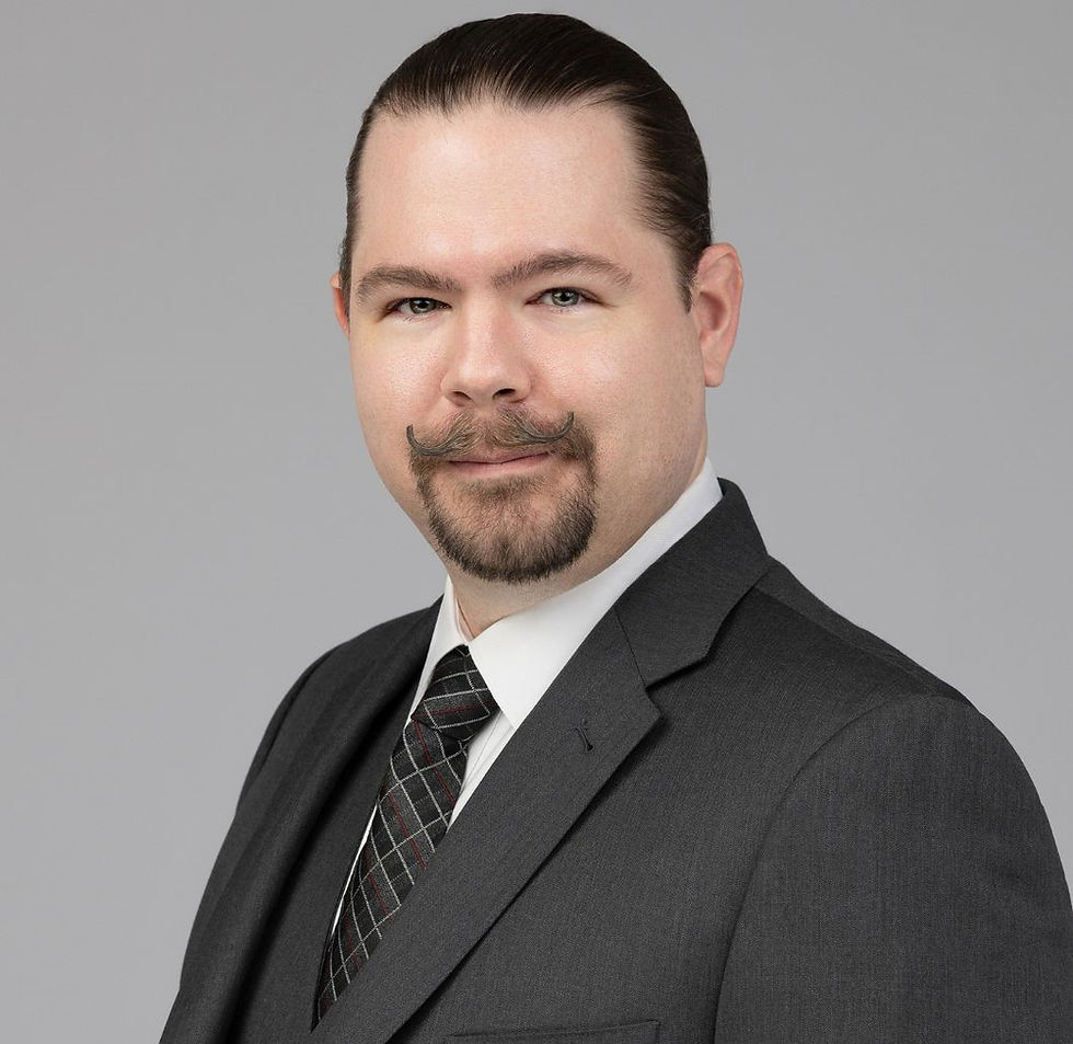 Man in a suit with a plaid tie and styled mustache, smiling subtly against a grey background. Professional and composed look.