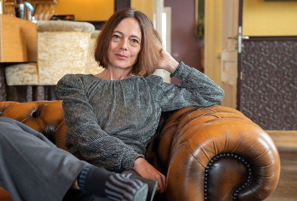 Woman with short brown hair sits relaxed on a brown leather couch, hand to head, wearing a gray blouse. Cozy indoor setting.