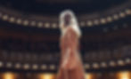 A woman in a floral gown stands on a stage in a grand theater. The audience watches, with warm lights creating an elegant atmosphere.