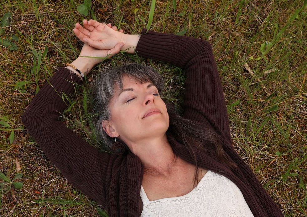 Woman with relaxed expression lies on grass, arms behind head, in a brown sweater and white lace top. Peaceful outdoor setting.