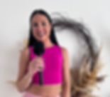 A person smiles, holding a black microphone against a white wall. They're wearing a bright pink top. A wreath with dried plants is in the background.