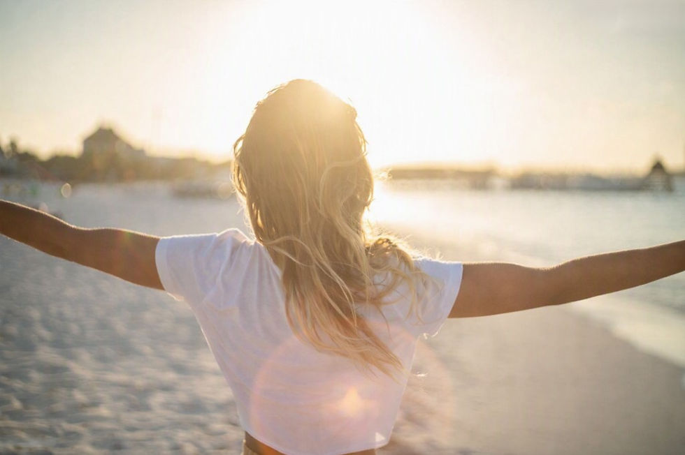 Woman in a white shirt stands on a beach with arms outstretched at sunset, glowing in warm golden light.