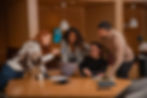 Four people engaged in discussion around a laptop in a cozy office. One holds a pen, others smile, with notebooks and coffee nearby. Warm lighting.
