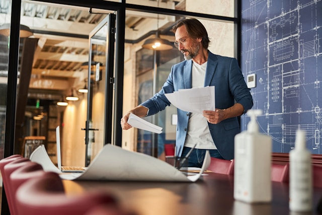 A man in a modern office is reviewing architectural plans and documents on a conference table.