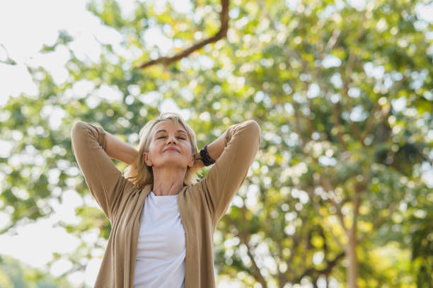 Woman relaxing with eyes closed and hands behind head in a sunny park. She's wearing a beige sweater, white top, and appears peaceful.