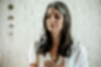 Woman with closed eyes holds incense, wearing white shirt and necklace. Background: white brick wall with hanging decor. Calm mood.