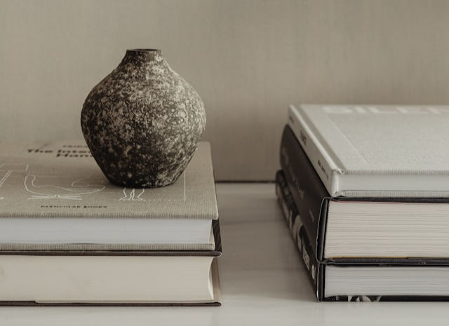 A speckled dark gray vase sits atop a stack of books next to another stack of books on a white surface.
