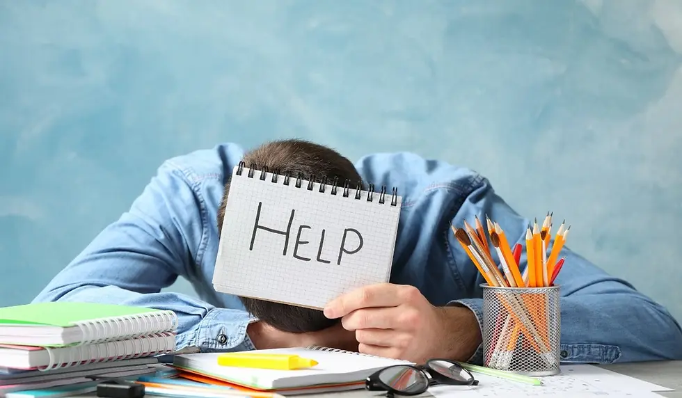 Person in denim shirt rests head on table with notebooks, holding a "Help" sign. Pencils in a holder; background is light blue.