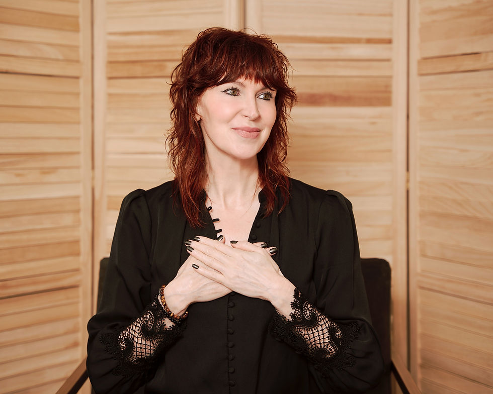 Red-haired woman in a black blouse smiles with hands on her chest against a light wood-paneled backdrop.