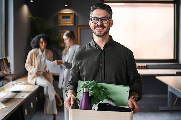 Smiling man in glasses holds a box with a plant and office supplies in a modern office, with coworkers blurred behind him.