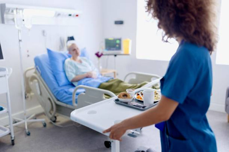 Nurse in blue uniform brings breakfast tray to patient in hospital bed. Room is bright, with medical equipment and a calm atmosphere.