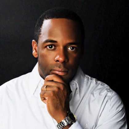 Man in a white shirt with a thoughtful expression rests his chin on hand. Dark background, wearing a watch and a wedding ring.