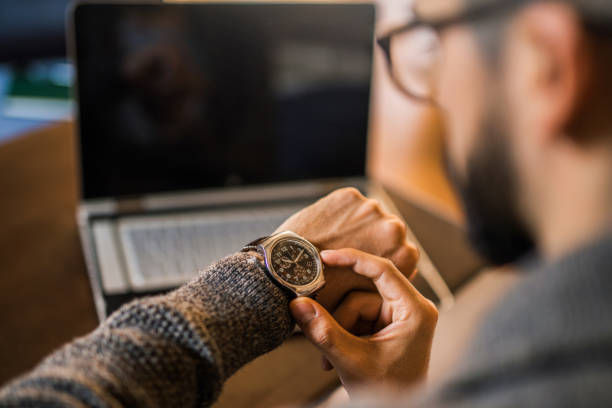 Man checking a watch on his wrist, seated near a laptop. Focus on the watch, with a blurred background. Cozy mood with warm tones.