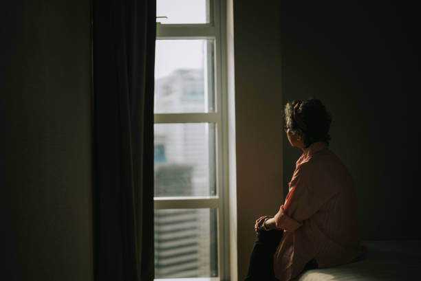 A woman sits on a bed, gazing out a window at a cityscape. The room is dimly lit, creating a contemplative mood.