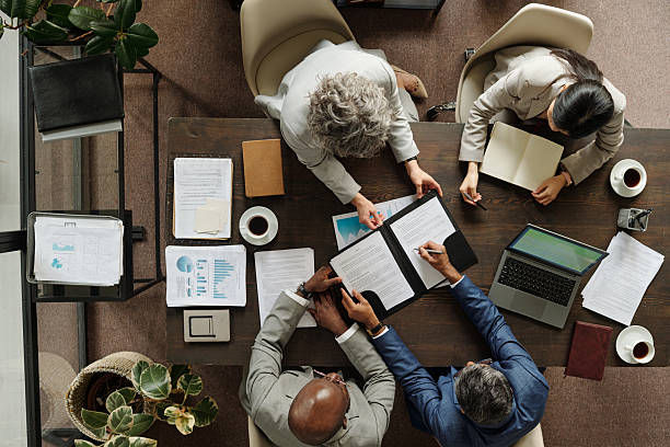 Top-down view of four coworkers reviewing documents around a conference table with a laptop, coffee cups, and plants.