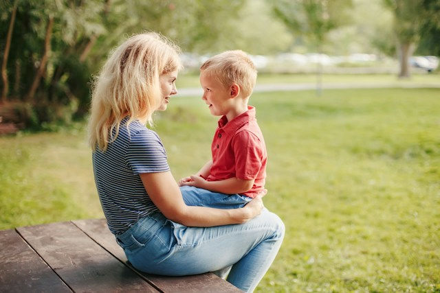 Blonde woman sits on a wooden bench holding a smiling young boy in a sunny park with green grass and trees.