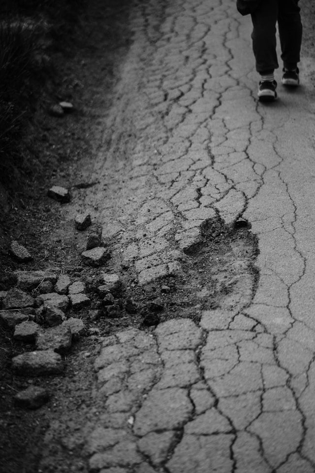 Cracked pavement with scattered debris. A person walks, partially visible. Black and white tones create a somber mood.