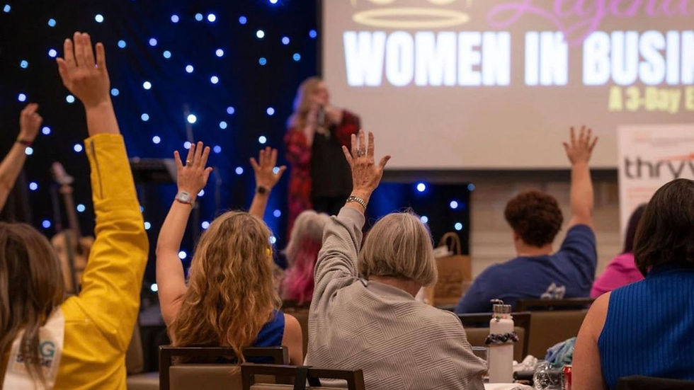 Women at a business event raise hands towards a speaker on stage. Background shows a screen with "Women in Business" text, starry lights.