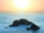 Rocks in a misty ocean at sunset, with an orange and blue sky. The scene is serene and peaceful with soft colors. No visible text.