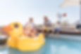 Family and kids relax by a desert pool on a giant yellow duck float, with a windmill and chalkboard sign in the background.