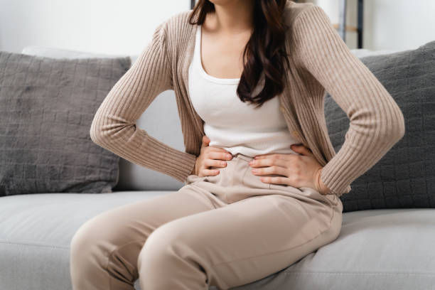 Woman in beige clothes sits on a gray couch, holding her stomach as if in pain. Neutral background. Mood suggests discomfort.