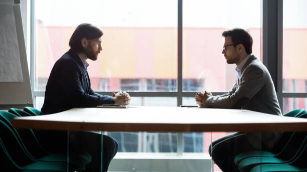 Two men in suits sit across a table in a modern office, engaged in a serious discussion. Large windows show a building outside.