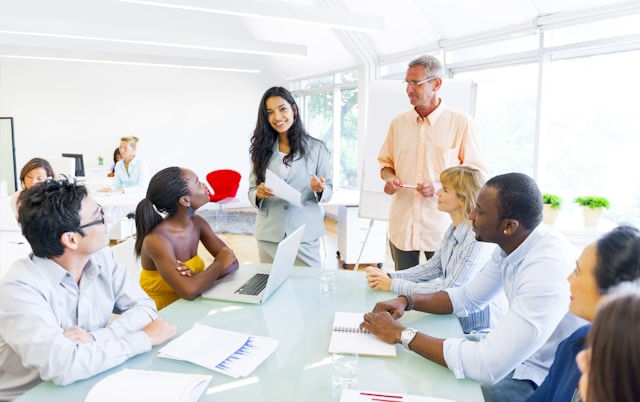 Professionals in a bright office engage in a meeting around a table, with one woman standing and speaking, and others listening attentively.