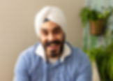 Man in white turban and striped shirt smiling, sitting in a room with green plants. Bright, cheerful atmosphere.