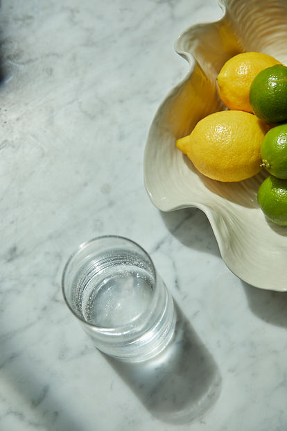 Glass of sparkling water on a marble surface, next to a shell-shaped bowl with lemons and limes.