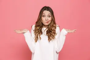 Woman in white sweater shrugging against a pink background, expressing confusion or uncertainty. Brown hair, casual pose.