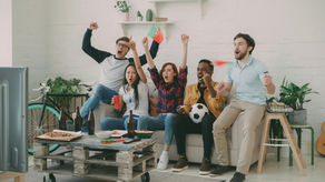 Friends cheer on a soccer match in a bright living room, waving flags and celebrating on a couch with snacks and drinks.