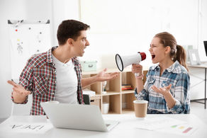 A woman in plaid shouts through a megaphone at a man in a checkered shirt, both gesturing angrily at a desk with a laptop and cups.