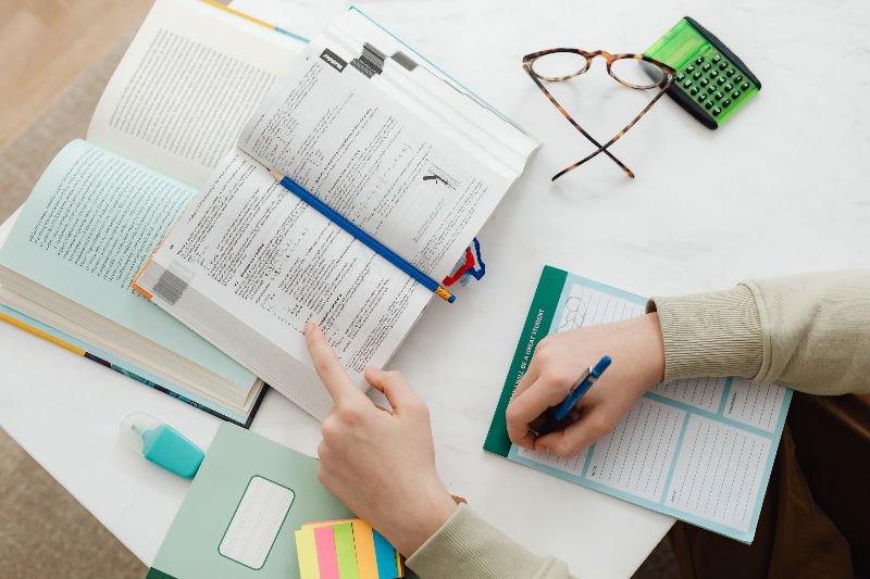 Person studying at a table with open books, a notebook, and a calculator. A hand writes notes. Glasses and sticky notes nearby. Bright setting.