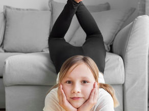 Petite fille blonde portant un tee shirt blanc et un legging noir qui regarde face à elle et qui se soutient la tête avec les deux mains sous le menton pendant que ses jambes sont sur le canapé.