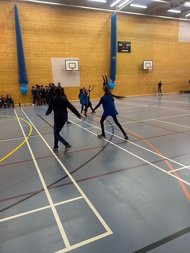 Fencing bout in a gymnasium, with two fencers in blue jackets on a court and spectators seated against the wall