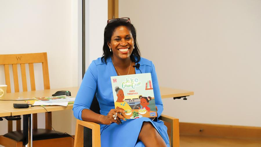 Woman in a blue dress smiling and holding a children’s book while seated in a classroom.