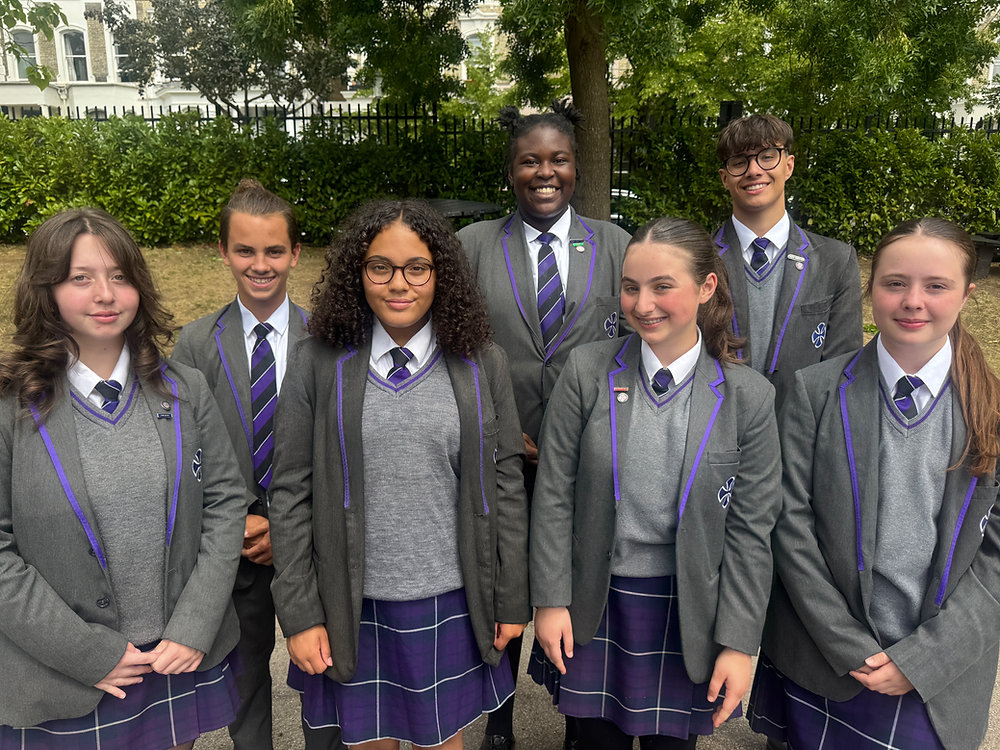 Six students in gray school uniforms stand outdoors in front of trees and a building.